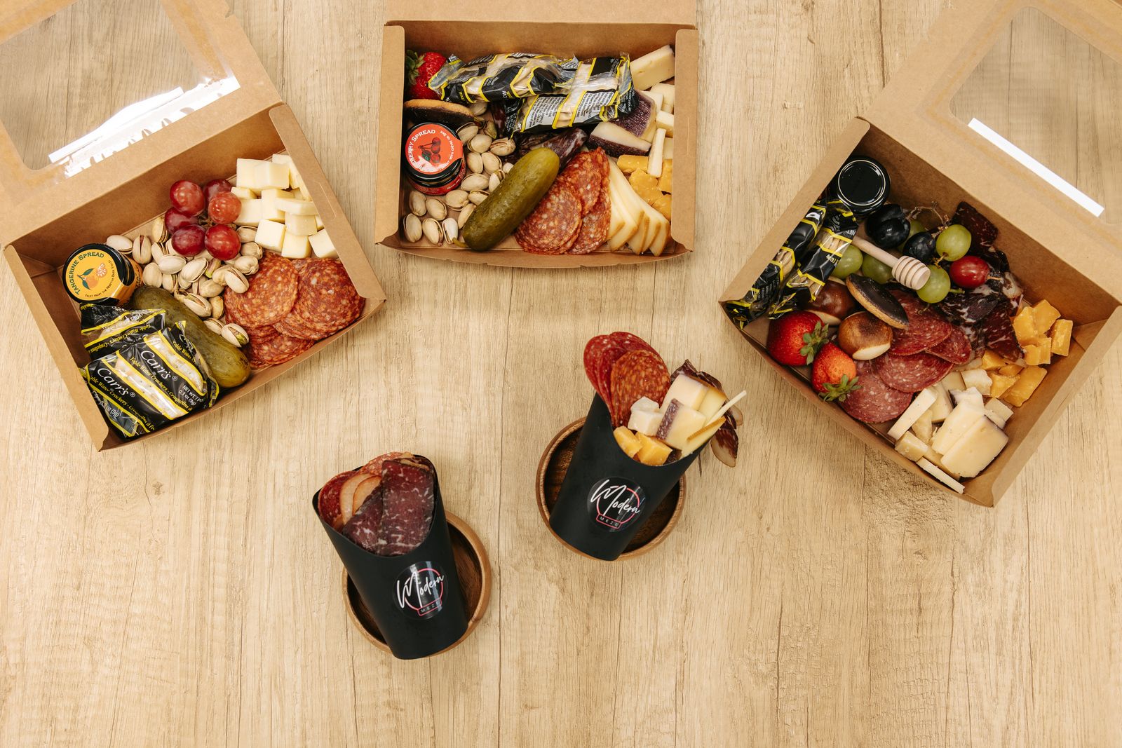 Three build-your-own charcuterie boxes and two meze cups on a wooden table
